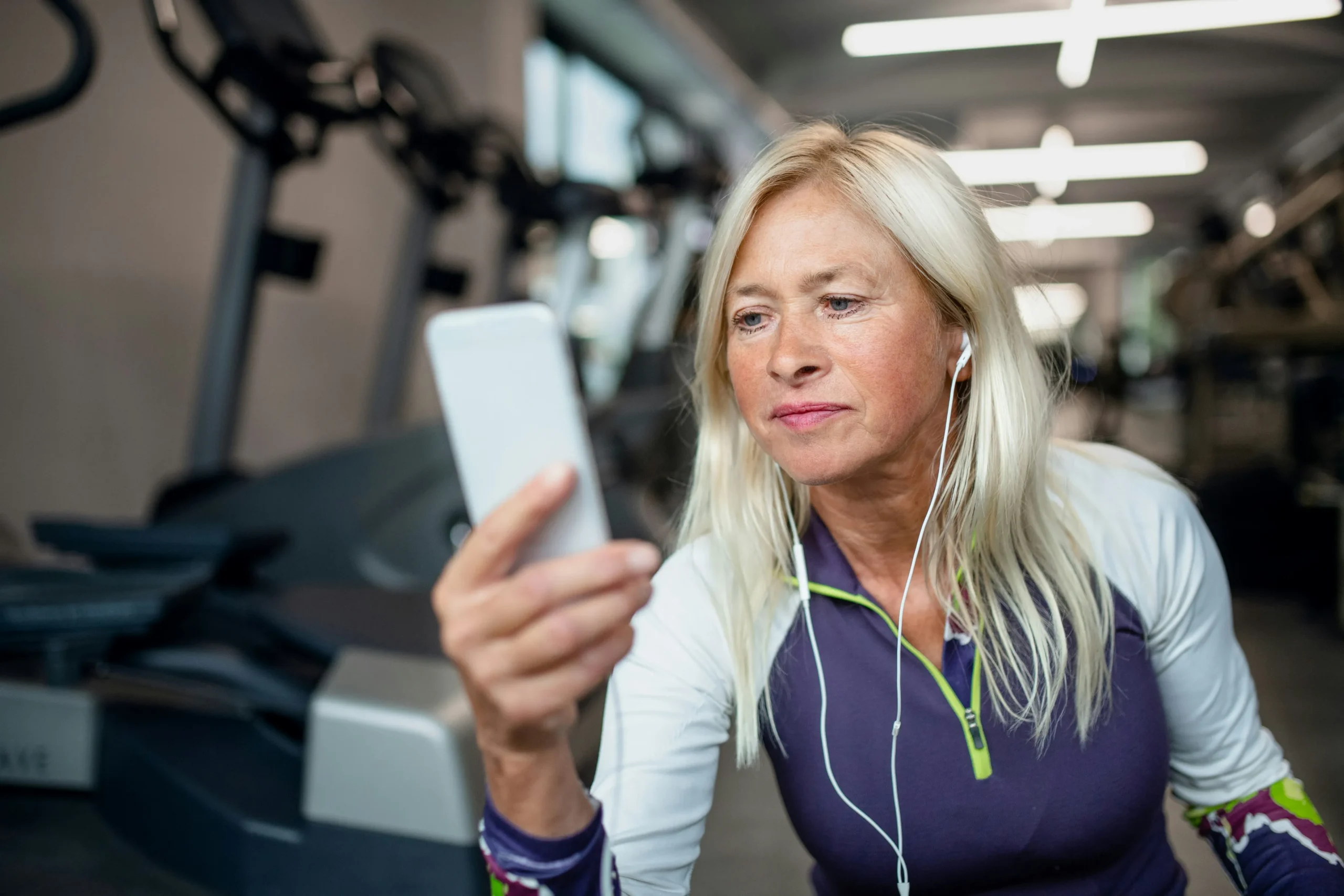 Middle-aged woman with blonde hair exercising in a gym, wearing earbuds and holding a smartphone to track her fitness, with gym equipment visible in the blurred background