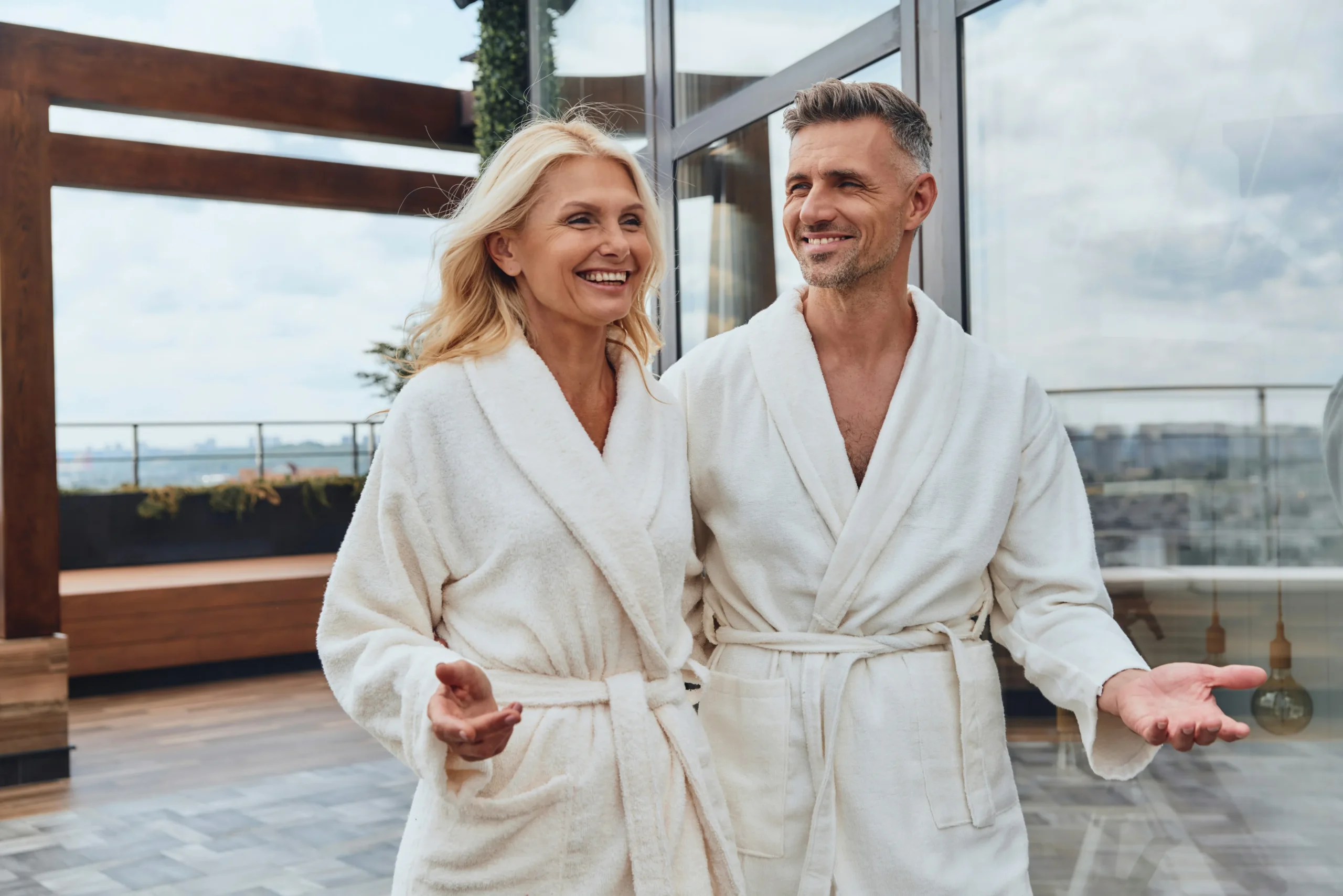Middle-aged couple wearing matching cream-colored robes, smiling and relaxing together on a modern terrace overlooking a scenic waterfront landscape with contemporary architecture